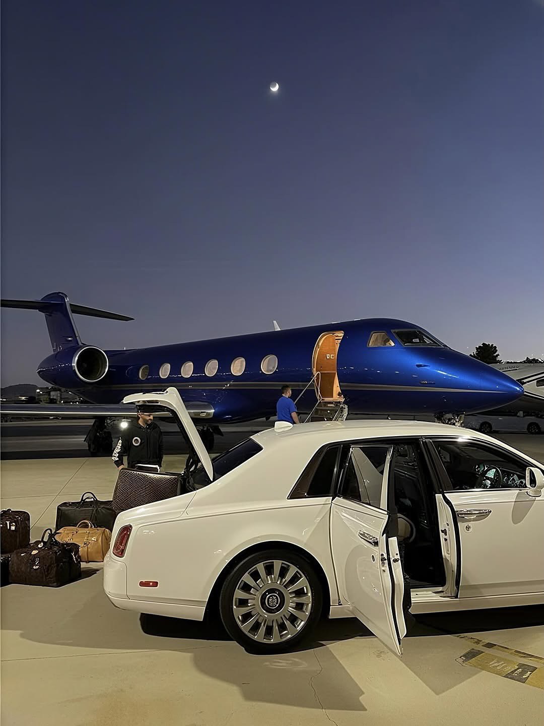 White luxury car and luggage beside a blue private jet at dusk